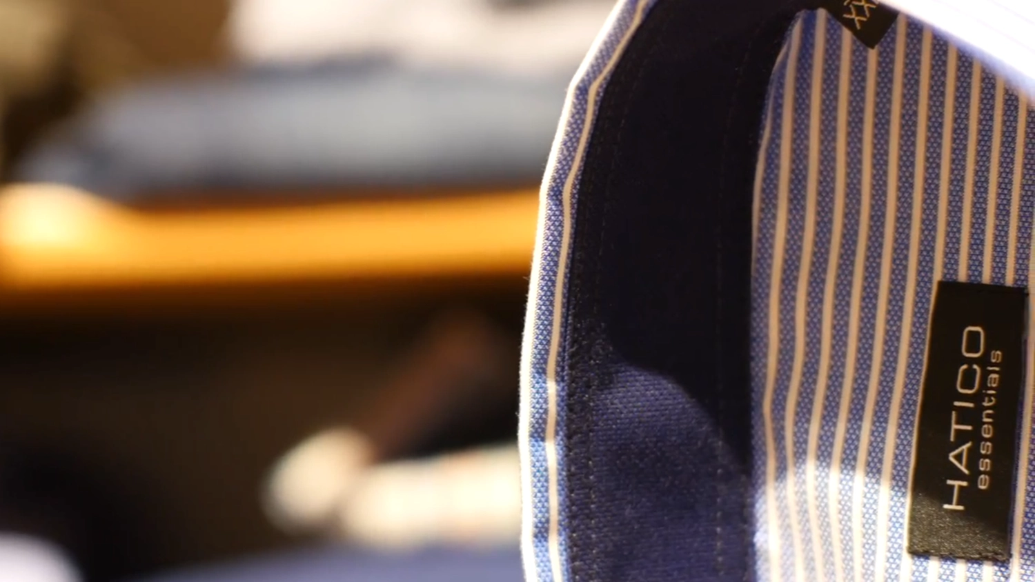 noBody´s perfect - Persönliche Betreuung, Maßanfertigungen und Bekleidung - Eine Nahaufnahme zeigt den Krageninnenseite und das Etikett eines blau-weiß gestreiften Hatico Essentials-Hemdes. Ein dunkelblauer Stoff füttert den Krageninnenseite und bildet einen Kontrast zu den hellen Streifen.