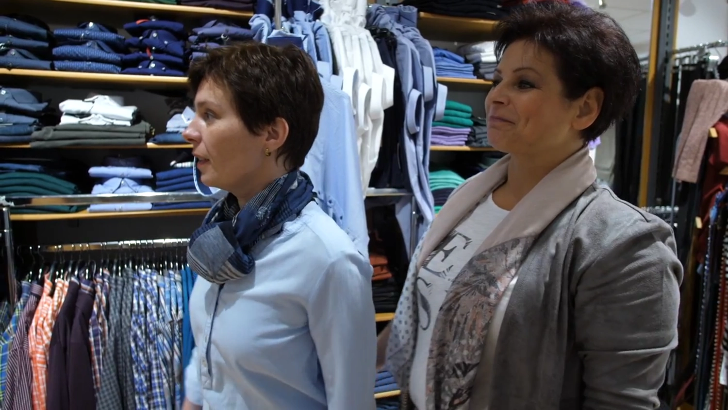 noBody´s perfect - Persönliche Betreuung, Maßanfertigungen und Bekleidung - Zwei Frauen sind in einem Bekleidungsgeschäft zu sehen. Die eine steht im Vordergrund und trägt ein hellblaues Hemd und einen gemusterten Schal. Die andere steht etwas hinter ihr rechts und trägt eine graue Jacke über einem weißen T-Shirt. Sie sind umgeben von Regalen und Kleiderstangen mit ordentlich gefalteten und hängenden Hemden in verschiedenen Farben.