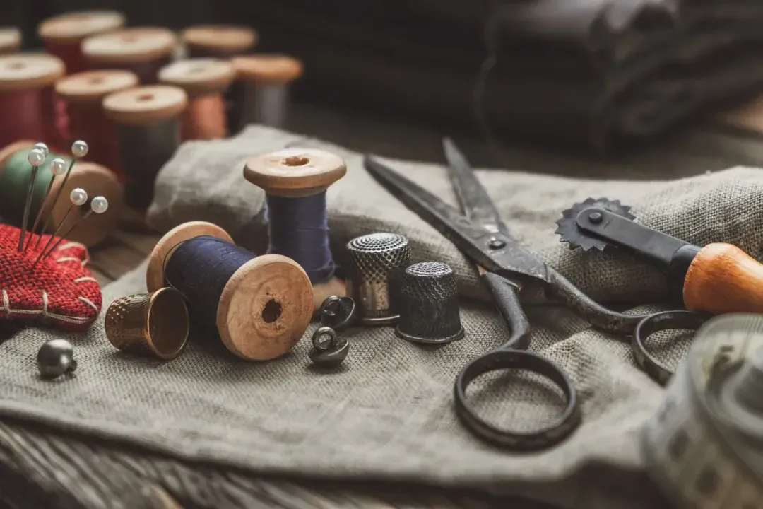noBody´s perfect - Persönliche Betreuung, Maßanfertigungen und Bekleidung - Auf einem Stück rustikalem Leinenstoff liegt eine Auswahl an Vintage-Nähutensilien, darunter hölzerne Garnspulen, Fingerhüte aus Metall, Scheren und ein Kopierrädchen.